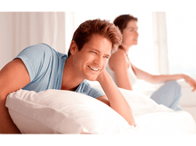 Charger l'image dans la galerie, Un homme à la peau claire sourit sur un lit, une femme est floue en arrière-plan.