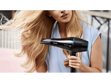 Charger l'image dans la galerie, Une femme sèche ses longs cheveux blonds avec un sèche-cheveux noir et bleu.