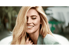 Charger l'image dans la galerie, Femme blonde souriante. Elle porte une veste verte et ses cheveux sont longs et ondulés. Le fond est flou.