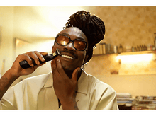 Charger l'image dans la galerie, Un homme noir se rase la barbe avec une tondeuse, souriant. Il porte des lunettes et une chemise claire.