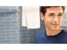 Charger l'image dans la galerie, Homme en chemise bleue, regardant la caméra, souriant. L'arrière-plan est un mur carrelé bleu et une serviette blanche.