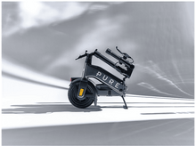 Charger l'image dans la galerie, Une trottinette électrique pliée sur fond blanc. La trottinette est gris foncé avec le mot 'PURE'.