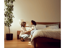 Charger l'image dans la galerie, Une femme et un enfant sont près d'un lit. La femme est accroupie et l'enfant est assis sur le lit. Une plante est à gauche.