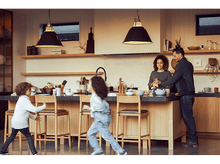 Charger l'image dans la galerie, Famille dans une cuisine moderne. Deux enfants courent près du comptoir. Les parents préparent de la nourriture derrière.