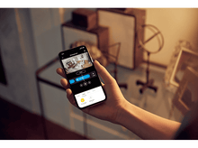 Charger l'image dans la galerie, Une main tient un téléphone affichant la vue d'une caméra de sécurité domestique. L'écran montre un salon. L'arrière-plan comprend de l'art et de la décoration.