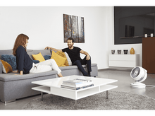 Charger l'image dans la galerie, Couple sur un canapé gris, table blanche avec des livres et un ventilateur blanc dans le salon.