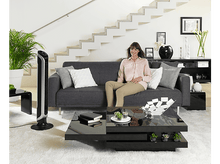 Charger l'image dans la galerie, Une femme assise sur un canapé gris. Un ventilateur à gauche, une table basse devant et un escalier.