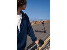 Charger l'image dans la galerie, Une personne avec des lunettes de soleil et une veste bleue se tient près d'un scooter et tient une tasse de café, avec la mer et le ciel en arrière-plan.