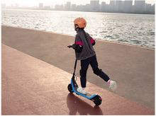 Charger l'image dans la galerie, Une fille sur un scooter bleu et noir sur une surface rouge, avec une skyline en arrière-plan.