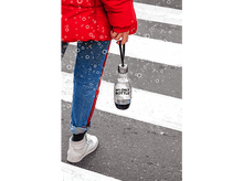 Charger l'image dans la galerie, Personne en veste rouge, jean bleu et chaussures blanches marche sur passage piéton, tenant bouteille.