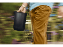 Charger l'image dans la galerie, Une personne tenant un haut-parleur portable noir marchant sur un arrière-plan flou de verdure.