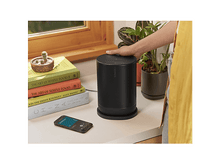 Charger l'image dans la galerie, Haut-parleur noir, livres et téléphone sur une surface blanche. Une main touche le haut-parleur.