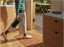 Charger l'image dans la galerie, Personne en pull vert et pantalon blanc traversant une porte, tenant un cylindre noir.