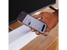 Charger l'image dans la galerie, Une main tient un téléphone affichant une interface de maison intelligente, avec un haut-parleur blanc au premier plan et une TV en arrière-plan.