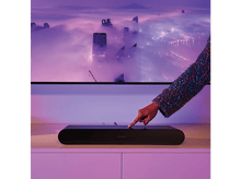 Charger l'image dans la galerie, Main touchant une barre de son Sonos noire sur une surface blanche. Fond flou avec paysage urbain.