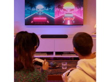 Charger l'image dans la galerie, Deux personnes jouant à des jeux vidéo sur un canapé. Une télévision affiche des graphismes de jeu.