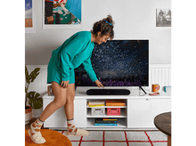 Charger l'image dans la galerie, Femme pointant un barre de son sur un meuble blanc. TV avec confettis derrière.