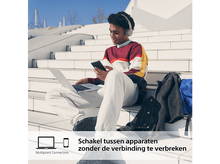 Charger l'image dans la galerie, Une personne assise sur des escaliers utilisant un ordinateur portable et un téléphone, portant un casque.