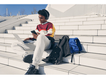 Charger l'image dans la galerie, Jeune homme assis sur des escaliers, utilisant un téléphone et un ordinateur portable. Sac à dos, casque et veste à proximité.