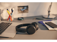 Charger l'image dans la galerie, Casque Sony noir sur un bureau avec un ordinateur portable, des stylos et une photo en arrière-plan.