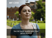 Charger l'image dans la galerie, Femme avec écouteurs blancs dehors. Arrière-plan: bâtiment et arbres.