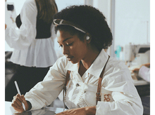 Charger l'image dans la galerie, Une femme travaille sur une tablette, portant un bandeau et tenant un stylet, dans un atelier.