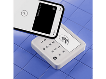 Charger l'image dans la galerie, Smartphone à écran noir au-dessus d'un terminal de paiement blanc sur une surface bleue.