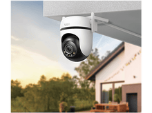 Charger l'image dans la galerie, Caméra Tapo montée sous un toit. Maison et arbres flous en arrière-plan au coucher du soleil.