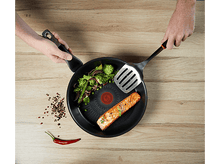 Charger l'image dans la galerie, Deux mains tenant une poêle avec nourriture et une spatule. Table en bois.