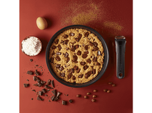 Charger l'image dans la galerie, Un grand cookie aux pépites de chocolat dans une poêle noire avec des ingrédients éparpillés.