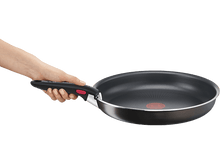 Charger l'image dans la galerie, Une main tient une poêle noire avec manche rouge et bord argenté. Fond noir.