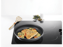 Charger l'image dans la galerie, Scène de cuisine : Filet de saumon dans une poêle, avec des légumes, sur une plaque de cuisson noire.