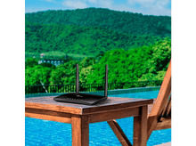 Charger l'image dans la galerie, Un routeur noir est posé sur une table en bois avec deux antennes. L'arrière-plan est une piscine et des montagnes.