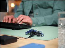 Charger l'image dans la galerie, Personne chargeant des piles avec un chargeur sur un tapis vert, avec un ordinateur portable en arrière-plan.