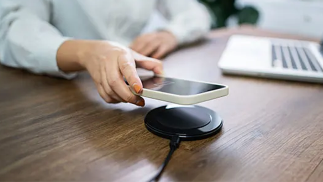 Une personne pose son smartphone sur un chargeur sans fil se trouvant sur un bureau. En arrière plan, on distingue les contours flous d'un pc portable.