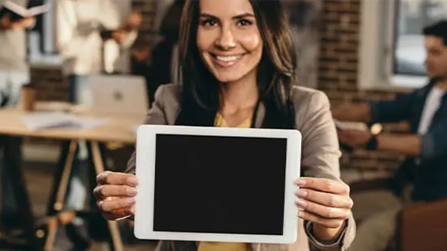 Une jeu femme souriante tient dans ses mains une tablette éteinte dans un open space.