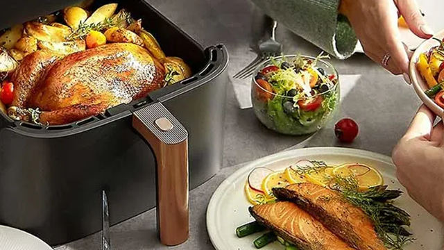 Un airfryer posé sur une table. La cuve est ouverte avec un poulet rôti garni de pomme de terres grillées à 'lintérieur.