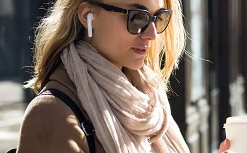 Une femme tient un téléphone et une tasse à café blanche à la main dans une rue ensoleillé. Elle porte des écouteurs aux oreilles.