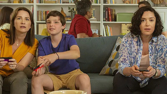 Deux femmes et un garçon sont assis sur un canapé et joue à la Nintendo Switch. En arrière plan deux hommes discutent.