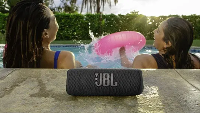 Une enceinte JBL sur le bord d'une piscine. En arrière blanc deux personnes rient aux éclats dans la piscine.
