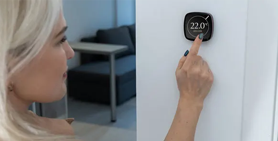 Une femme règle la température d'un thermostat connecté affichant 22 °C sur le mur d'une maison.