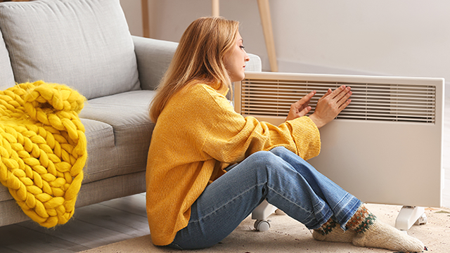 Une femme habillé en jaune est assise au sol et se réchauffe près d'un radiateur électrique à côté d'un canapé.