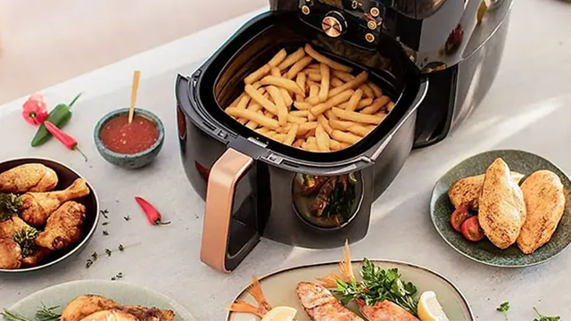 Un airfryer dont la cuve ouverte est rempli de frites. Il est posé sur une table ou d'autres plats sont présents.