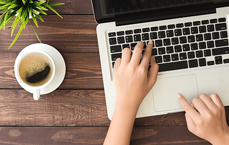 Une vue de dessus de mains tapant sur le clavier d'un pc portable. Une tasse de café est également posé sur le bureau.