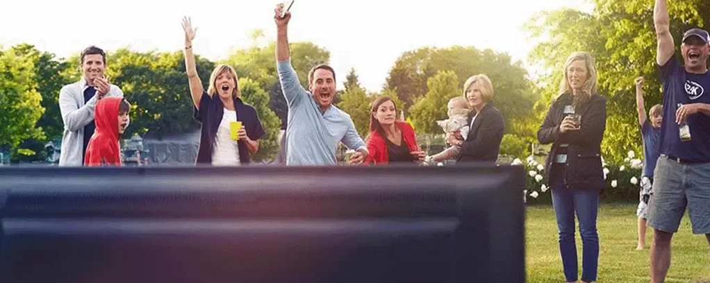 Un groupe de personne regarde la télévision dans un jardin avec des verres à la main.