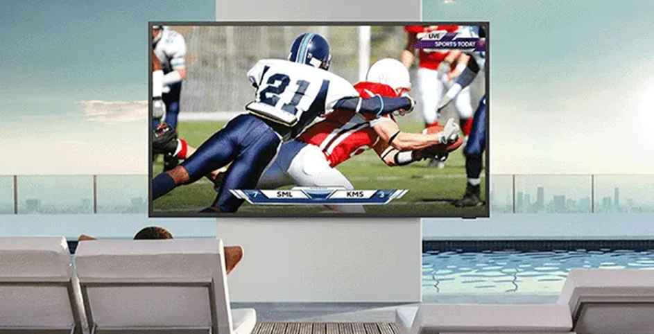 Une personne couché sur un transat regarde un match de football américain sur une TV installée au bord d'une piscine.