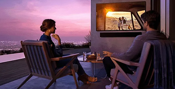 Un couple regarde un film sur un téléviseur installé en terrasse avec vue sur un coucher de soleil.