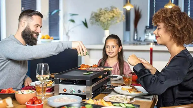 Une famille souriante autour d'un appareil à pierrade prend son repas avec  assiettes de viandes et légumes.