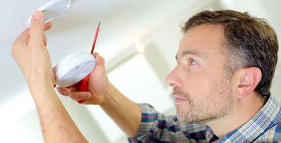 Un homme fixe un détecteur de fumée au plafond avec un tournevis.