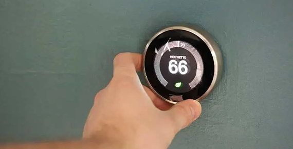Une main règle un thermostat connecté fixé au mur et affichant une température de 66° F.